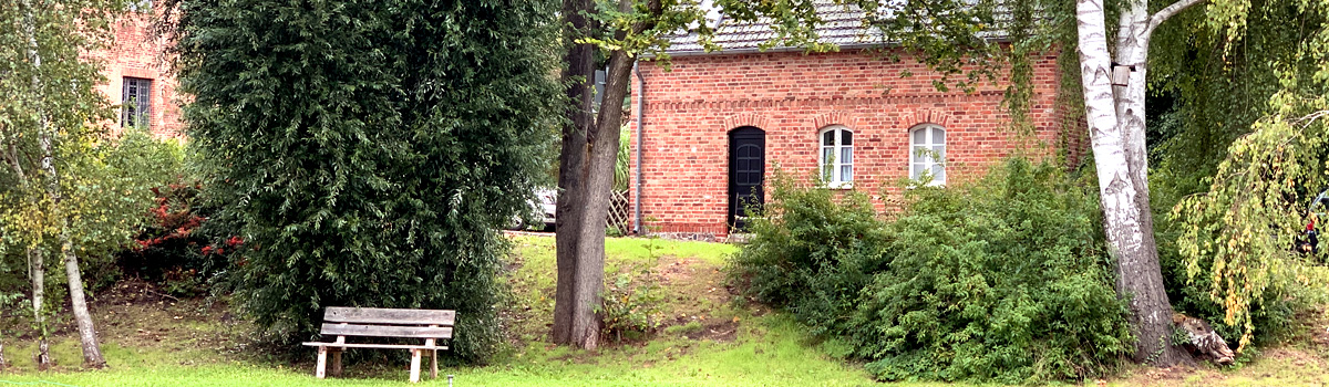 Ein Backsteingebäude hinter verschiedensten Baumarten und einer Sitzbank. 